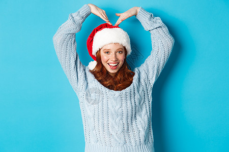 寒假和平安夜的概念 穿着圣诞帽和毛衣的可爱红发少女 做着心形手势 微笑着 祝圣诞快乐 站在蓝色背景上 舒适 促销图片
