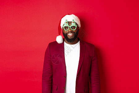 新年广告戴着圣诞帽和派对眼镜的风趣黑人庆祝圣诞节 微笑着开心地祝圣诞快乐 站在红色背景上 工作室 庆典背景