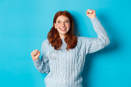 快乐的红发女孩为团队扎根 用举起手欢呼 微笑 庆祝胜利或节日 站在蓝背景下站立 促销 新年图片