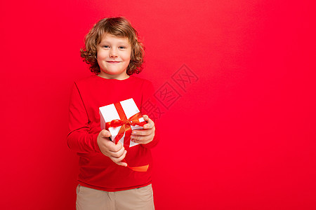 照片中 快乐微笑的金发卷发男孩被隔离在红色背景墙上 身穿红色毛衣 拿着礼盒 看着相机 孩子 男生图片