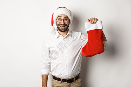 圣诞快乐 假期概念 戴着圣诞帽的兴奋的大胡子男人拿着圣诞袜 微笑着庆祝新年 白色背景 幸福图片