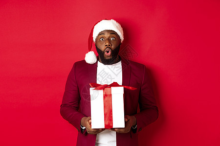 圣诞 新年和购物概念 奇异的非洲男子在圣塔帽子上说哇 拿着节日礼物看摄影机 惊讶地看到红色背景 假期 冬天图片