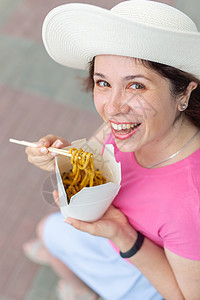 身戴帽子的年轻女子在温暖的夏天在公园散步时吃中国面条 外送食物概念 笑声 快乐 时髦图片