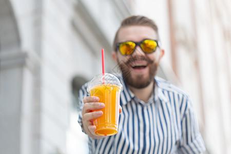 一个模糊的时尚年轻时髦男性的肖像 留着胡子和小胡子 手里拿着新鲜的橙汁 健康零食的概念 眼镜 城市图片