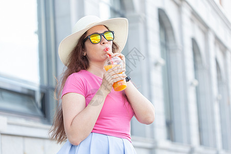 在阳光明媚温暖的夏日 一位戴着帽子和眼镜 手里拿着果汁在城市里走来走去的开朗积极的年轻女子的画像 无忧无虑的心情的概念 青年图片