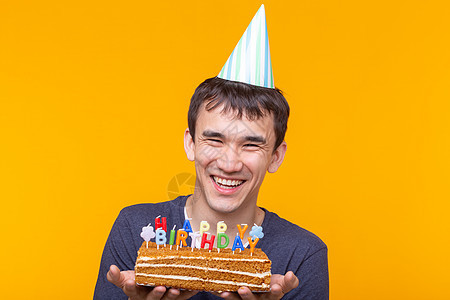 积极有趣的亚洲年轻男子戴着帽子 手里拿着燃烧的蜡烛和自制蛋糕 背景是黄色的 周年纪念和生日概念 纸杯蛋糕 美丽的图片