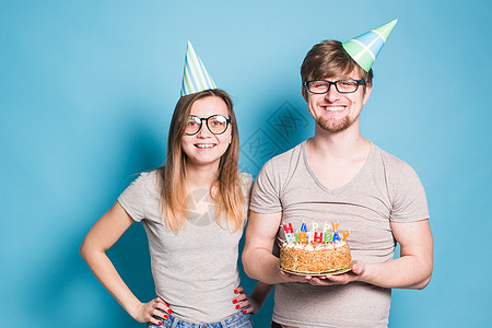 有趣的书呆子男人和女人都戴着节日帽和眼镜 手里拿着蓝色背景上点着蜡烛的生日蛋糕 喜悦 笑图片
