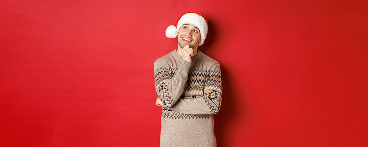 戴着圣诞帽和圣诞毛衣的快乐年轻人的形象 想象着一些东西 想着新年礼物 微笑着 看着左上角 站在红色背景上 派对 庆典背景图片