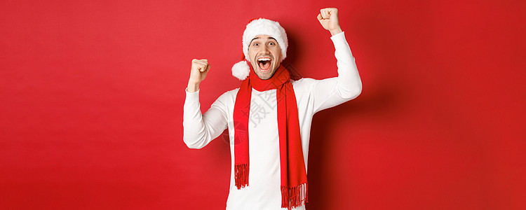 欢乐的男子在圣塔帽和围巾中 呼喊欢乐 举起手 庆祝胜利或胜利 战胜红色背景 新年 派对图片