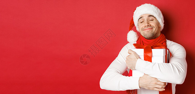 寒假 圣诞节和生活方式的概念 戴着圣诞帽和围巾的可爱男人的形象 拥抱他的新年礼物 微笑着受宠若惊 站在红色背景上 购物 情感背景图片