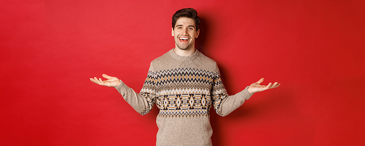 庆祝新年假期的快乐帅哥的画像 穿着圣诞毛衣 双手横向展开 微笑着 拿着一些东西在复印空间 站在红色背景上 快乐的 广告背景图片