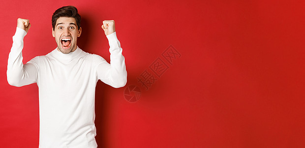 穿着白色毛衣 举起手来 胜利 庆祝新年 在红色背景下欢乐的幸运男子的喜悦肖像 情感 圣诞节图片