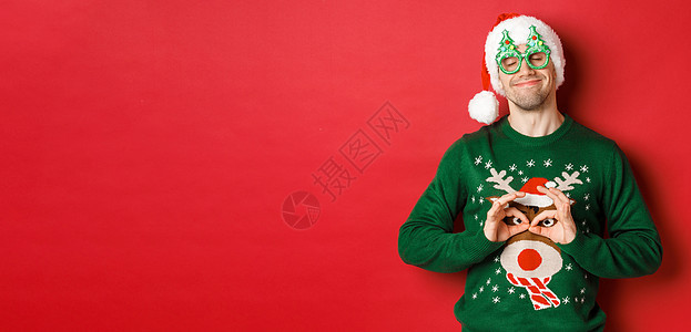 戴着派对眼镜和圣诞老人帽子的快乐微笑男子的形象 穿着滑稽的圣诞毛衣四处闲逛 庆祝寒假 站在红色背景上 广告 圣诞节图片