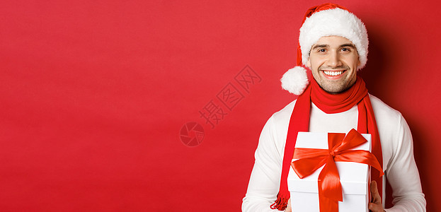 寒假 圣诞节和生活方式的概念 戴着圣诞帽和围巾 拿着礼物 送新年礼物和微笑 红色背景的留着胡须的帅哥的特写 时尚 庆典背景图片