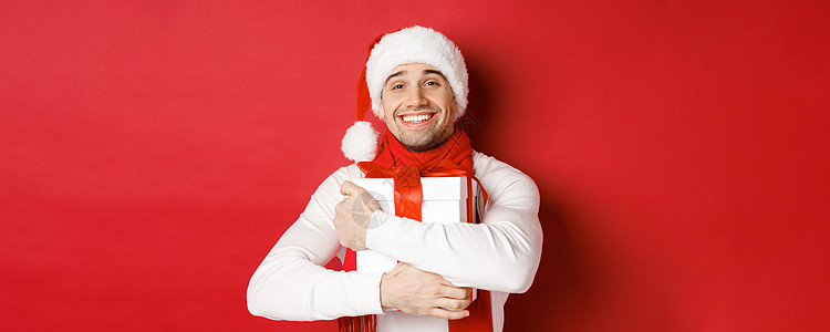 寒假 圣诞节和生活方式的概念 戴着圣诞帽和围巾的可爱男人的形象 拥抱他的新年礼物 微笑着受宠若惊 站在红色背景上 冬天 快乐的背景图片