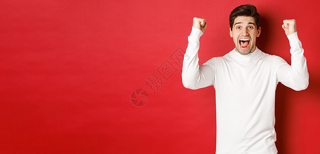 穿着白色毛衣 举起手来 胜利 庆祝新年 在红色背景下欢乐的幸运男子的喜悦肖像 黑发 成人图片