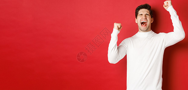 身穿白色毛衣的帅哥的画像 心情愉快 庆祝胜利 欢呼雀跃 举起双手迎接胜利 站在红色背景上 圣诞节 快乐的图片
