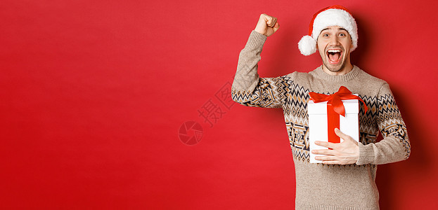 快乐而兴奋的帅哥的形象 reicing 圣诞礼物 胜利地举起手 微笑着 庆祝新年 站在红色背景上 戴着圣诞老人的帽子 成功 大胡图片