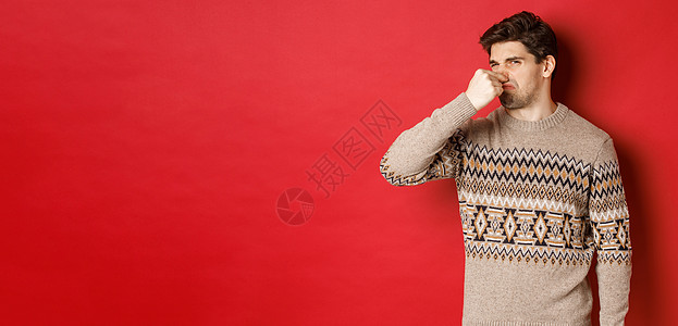 令人厌恶的年轻人在冬季毛衣上 闭上鼻子和严酷地从臭气熏天 站在红色背景上站立 成人 脸图片