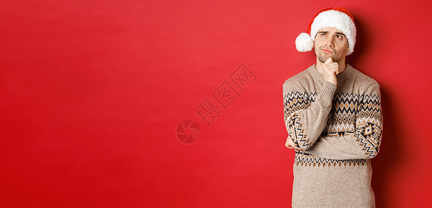 穿着圣诞毛衣和圣诞帽的帅哥形象 想着新年礼物 深思熟虑地看着左上角 站在红色背景上 快乐 微笑背景图片