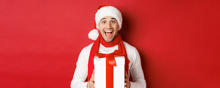寒假 圣诞节和生活方式的概念 身穿圣诞老人和围巾的帅哥兴奋的特写 看起来很惊讶 收到新年礼物 站在红色背景上 成人 男生背景图片