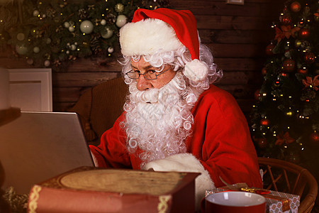 圣诞老人用笔记本电脑坐在壁炉附近的舒适房间 信 写作图片
