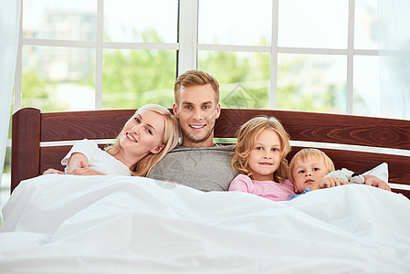 家庭日 年轻美丽的四口之家一起躺在床上放松 微笑着看着相机 在家享受周末图片
