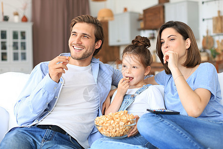 孩子坐在沙发上看电视 吃爆米花 年轻父母抱着女儿在沙发上一起放松 家庭快乐 幸福的家庭 老的图片