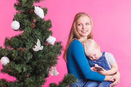 圣诞节和节日概念 — 粉红色背景中微笑的女人抱着小女儿靠近圣诞树的画像 冬天 快乐的图片