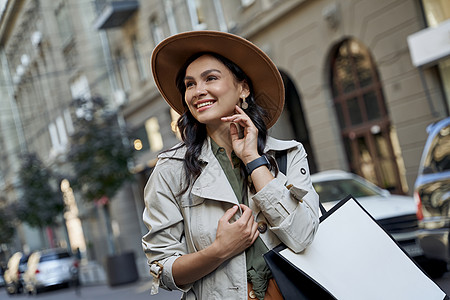 时尚女孩 一位戴着帽子 戴着购物袋的年轻漂亮时尚女性的画像站在城市街道上 一边看着一边微笑图片
