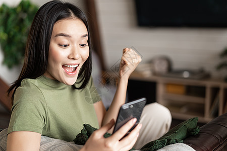 微笑的年轻女性看着满意 做拳手泵和阅读手机 坐在家里并取得胜利 房间 互联网图片
