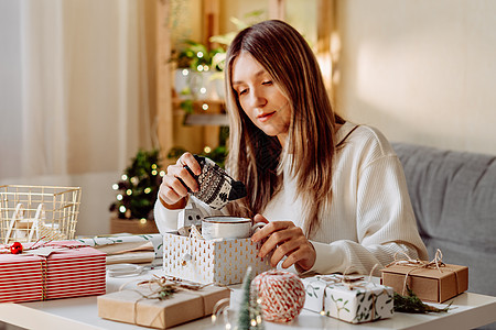 女人在家里包装圣诞生态天然礼品盒 在白桌上准备礼物 包括装饰元素 灯光和圣诞或新年 DIY 包装概念 冷杉 冬天图片