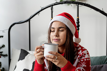 穿着红色毛衣和圣塔帽的年轻女士坐在床上庆祝圣诞节 喝杯热饮 美丽的 家图片