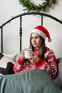 穿着红色毛衣和圣塔帽的年轻女士坐在床上庆祝圣诞节 喝杯热饮 礼物 幸福图片