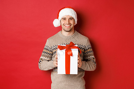 身着圣诞帽和冬季毛衣的英俊微笑男子的形象 拿着礼物 送圣诞礼物 祝节日快乐 站在红色背景上 美丽的 男生图片
