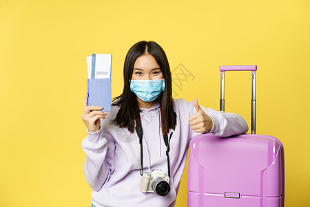 戴医用面具的日本女孩 出示护照和机票 竖起大拇指 带着手提箱摆姿势 去度假 黄色背景图片