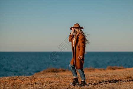 一个女人沿着海边的海岸散步 一位身穿棕色外套 戴着时尚妆容的帽子的优雅女士在海边散步 水 闲暇图片