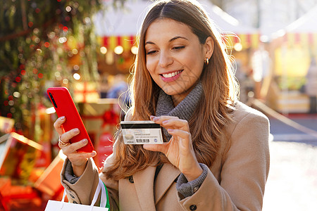 美丽优雅的女人看着信用卡号码插入智能手机在平安夜进行网上购物 户外信用卡支付图片
