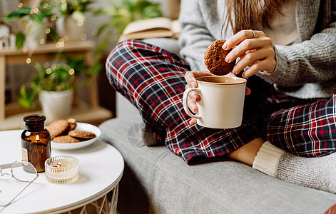 穿着针织冬季保暖袜子 毛衣和格子睡衣的舒适女人腿 坐在家里的沙发上 喝着热可可或杯子里的咖啡 与蜡烛 装饰 曲奇饼和室内植物的秋图片