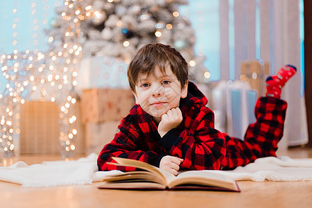 一个穿着睡衣的男孩在圣诞树下看书 新年的心情 在读书 儿童读物 礼物 床图片