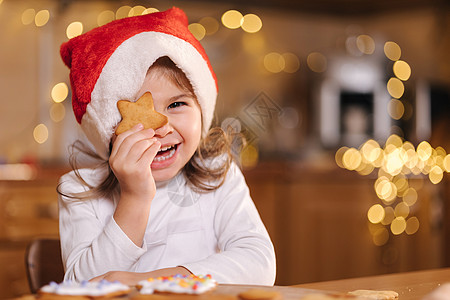 戴着圣诞老人帽子的可爱小女孩在姜饼旁闭上一只眼睛 笑着 快乐的小孩为圣诞节做准备 童话般的灯光散景背景 烹饪 面粉图片