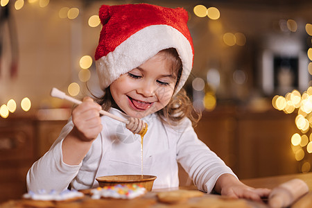 穿着红圣塔帽的小女孩用蜂棒吃蜂蜜 用童话灯装饰的厨房厨房 在家过圣诞节 新年快乐图片