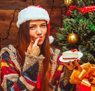 聪明的女士拿着一个盘子 里面放着一块蛋糕 嘴里放着手指 平安夜 圣诞晚餐 圣诞蛋糕 圣诞树 圣诞礼物 木头背景 特写图片