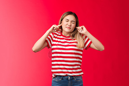 天哪可怕的噪音 心烦意乱和不高兴的金发韩国女孩做鬼脸不喜欢大声烦扰的声音塞手指耳孔不愿意听听废话 站在红色背景 学生 惊讶图片