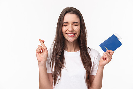 旅游 假期 夏季概念 旅途乞讨 穿着 T 恤 快乐开朗的黑发女孩 闭上眼睛 微笑 许愿 交叉手指祝你好运 拿着带机票的护照 商业图片