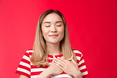 爱温暖的少女心 温柔浪漫的亚洲女人闭上眼睛感觉满足乐观 按手胸微笑放松愉快 记住美好的回忆 感受和平幸福红色背景 多样性 金发图片