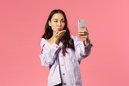 在线生活方式 人与美的概念 可爱的现代时尚亚洲女孩 网络博主通过手机摄像头发送亲吻 自拍或录制视频 粉红色背景 有趣的 健康图片