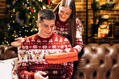 30 多岁的年轻女性遮住微笑好奇的丈夫的眼睛 在装饰过的节日树附近送上装有圣诞礼物的包装盒 新年寒假家庭庆祝活动 假日奇迹图片