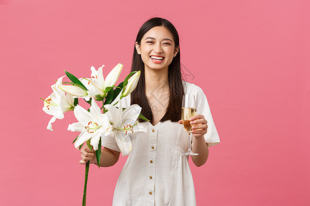 庆祝活动 派对假期和有趣的概念 穿着白色裙子的愚蠢的生日快乐女孩 在收到美丽的百合花束时笑得大大的 手里拿着香槟 站在粉红色的背图片