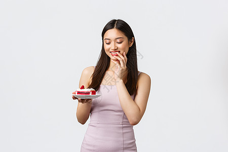 生活方式 假期 庆祝活动和食品概念 美丽的亚洲女孩咬手指 微笑着看着一块蛋糕 想吃甜美的甜点 诱人的节食作弊图片